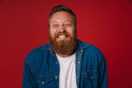 Téléchargez les photos : Adulte barbu tatoué beau roux tendu homme avec les yeux fermés et les dents serrées debout sur fond rouge isolé - en image libre de droit