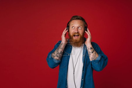Téléchargez les photos : Portrait d'adulte barbu tatoué bel homme rousse enthousiaste élégant avec bouche ouverte dans les écouteurs les touchant et regardant de côté, tout en se tenant sur fond rouge isolé - en image libre de droit