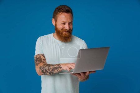 Foto de Adulto guapo pelirrojo barbudo tatuado sonriente hombre sosteniendo el ordenador portátil y escribiendo en él, mientras que de pie sobre fondo azul aislado - Imagen libre de derechos