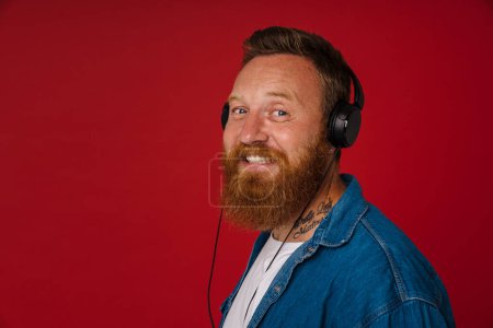 Téléchargez les photos : Portrait d'adulte barbu tatoué beau homme rousse souriant élégant dans les écouteurs regardant la caméra, tout en se tenant sur fond rouge isolé - en image libre de droit