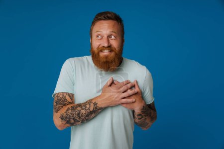 Téléchargez les photos : Adulte rousse barbu tatoué homme souriant en t-shirt avec les mains pliées sur la poitrine exprimant une émotion tendre et regardant de côté, tout en se tenant sur fond bleu isolé - en image libre de droit