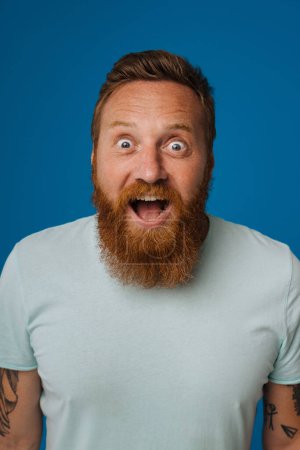 Téléchargez les photos : Adulte barbu tatoué bel homme élégant et enthousiaste avec la bouche ouverte, regardant la caméra, tout en se tenant sur fond bleu isolé - en image libre de droit