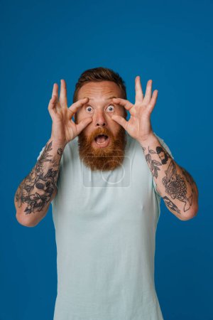 Foto de Retrato de adulto barbudo tatuado guapo hombre elegante con los ojos bien abiertos con las manos y mirando a la cámara, mientras que de pie sobre el fondo azul aislado - Imagen libre de derechos