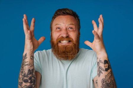Photo for Adult bearded tattooed handsome stylish stressed man with clenched teeth raised hands and splayed fingers looking upwards, while standing over isolated blue background - Royalty Free Image