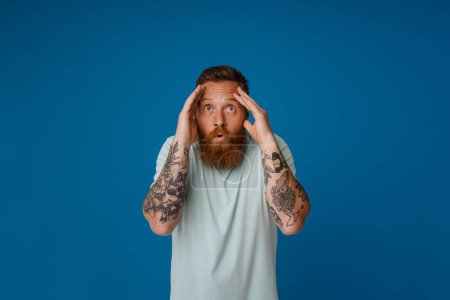 Photo for Adult bearded tattooed handsome stylish shocked man touching his head and looking aside, while standing over isolated blue background - Royalty Free Image