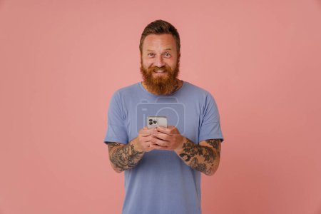 Téléchargez les photos : Portrait d'adulte barbu tatoué bel homme souriant tenant le téléphone et regardant la caméra, tout en se tenant sur fond rose isolé - en image libre de droit