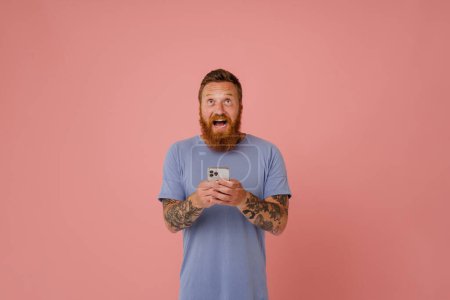 Téléchargez les photos : Portrait d'adulte barbu tatoué bel homme enthousiaste tenant son téléphone et regardant vers le haut, tout en se tenant sur fond rose isolé - en image libre de droit
