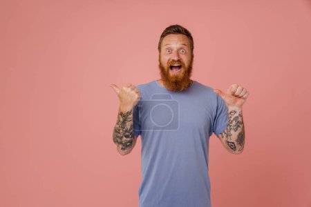 Photo for Adult handsome redhead bearded tattooed enthusiastic man with opened mouth pointing aside with thumbs both raised hands and looking at camera, while standing over isolated pink background - Royalty Free Image