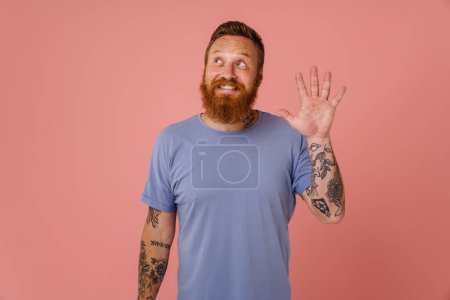 Téléchargez les photos : Adulte beau rousse barbu tatoué souriant heureux homme heureux agitant et regardant de côté tout en se tenant sur fond rose isolé - en image libre de droit