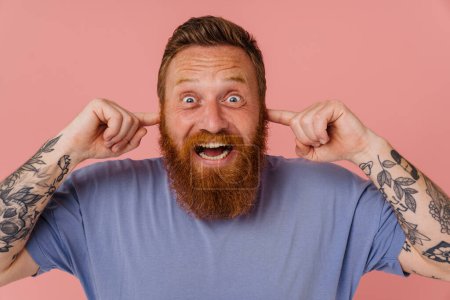 Téléchargez les photos : Adulte rousse barbu tatoué homme stressé en t-shirt avec bouche ouverte bouchant ses oreilles avec les doigts et regardant la caméra, tout en se tenant sur fond rose isolé - en image libre de droit