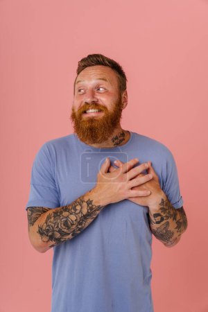 Téléchargez les photos : Adulte rousse barbu tatoué homme souriant en t-shirt avec les mains pliées sur la poitrine regardant de côté, tout en se tenant sur fond rose isolé - en image libre de droit