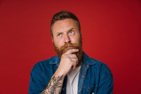 Photo for Ginger puzzled man with tattoo frowning and looking upward isolated over red background - Royalty Free Image