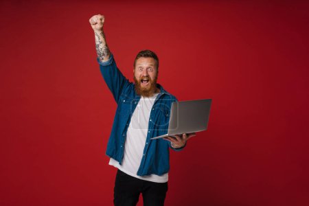 Téléchargez les photos : Ginger excité homme portant chemise en denim gestuelle et en utilisant ordinateur portable isolé sur fond rouge - en image libre de droit