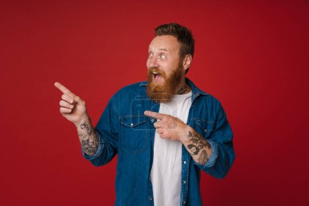 Téléchargez les photos : Gingembre excité homme avec tatouage geste et pointant les doigts de côté isolé sur fond rouge - en image libre de droit