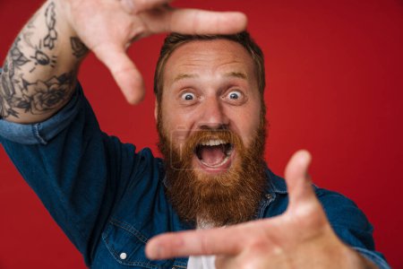 Téléchargez les photos : Gingembre choqué homme avec tatouage faisant geste de cadre isolé sur fond rouge - en image libre de droit