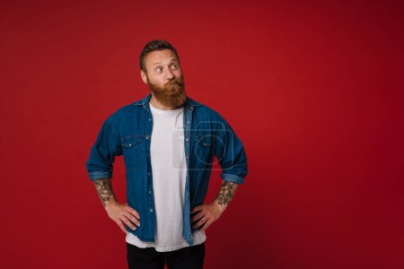 Photo for Ginger puzzled man with tattoo grimacing and looking aside isolated over red background - Royalty Free Image