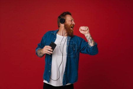 Photo for Ginger white man singing and dancing while listening music isolated over red background - Royalty Free Image