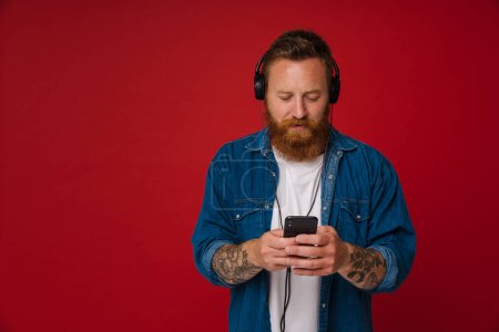 Téléchargez les photos : Ginger homme blanc dans les écouteurs écouter de la musique et en utilisant un téléphone cellulaire isolé sur fond rouge - en image libre de droit