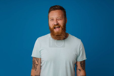 Téléchargez les photos : Gingembre excité homme avec tatouage clin d'oeil et sourire à la caméra isolé sur fond bleu - en image libre de droit