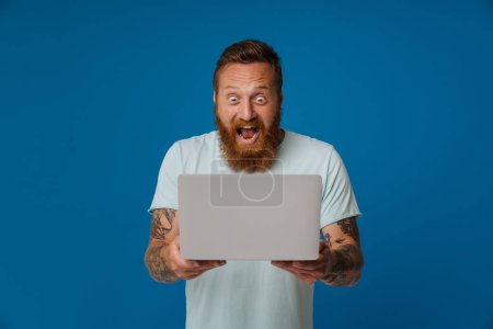 Téléchargez les photos : Gingembre excité homme avec tatouage crier et travailler avec ordinateur portable isolé sur fond bleu - en image libre de droit