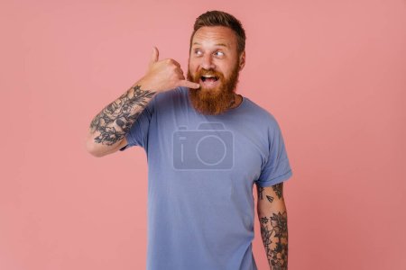 Foto de Hombre excitado jengibre con barba sonriendo y haciendo gesto de teléfono aislado sobre fondo rosa - Imagen libre de derechos