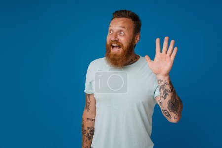 Téléchargez les photos : Ginger excité homme avec tatouage souriant et agitant la main à la caméra isolé sur fond bleu - en image libre de droit