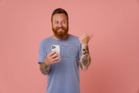 Photo for Ginger white man using cellphone and pointing finger aside isolated over pink background - Royalty Free Image