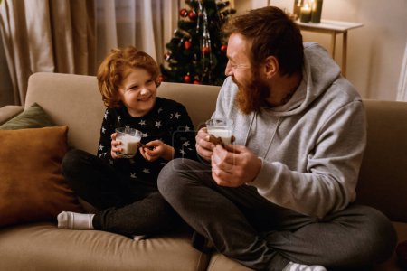 Foto de Alegre padre e hijo bebiendo leche con galletas mientras descansan en el sofá en la acogedora sala de estar con árbol de Navidad en el fondo - Imagen libre de derechos