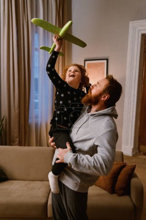 Téléchargez les photos : Père joyeux et son petit fils jouant avec l'avion jouet dans le salon confortable - en image libre de droit