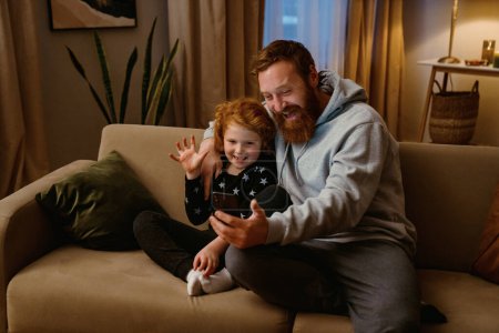 Téléchargez les photos : Père et fils excités ayant un appel vidéo via téléphone portable tout en étant assis sur le canapé dans le salon confortable - en image libre de droit