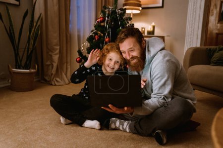 Téléchargez les photos : Gingembre joyeux père et fils ayant appel vidéo via ordinateur portable tout en étant assis dans le salon confortable avec arbre de Noël sur le fond - en image libre de droit