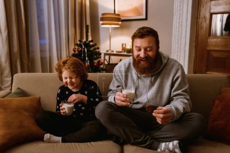 Foto de Padre e hijo pequeño bebiendo leche y comiendo galletas mientras están sentados en el sofá en la acogedora sala de estar con árbol de Navidad en el fondo - Imagen libre de derechos