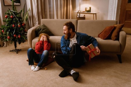 Foto de Lindo niño recibiendo regalo de su padre mientras está sentado en el suelo en la acogedora sala de estar con árbol de Navidad en el fondo - Imagen libre de derechos