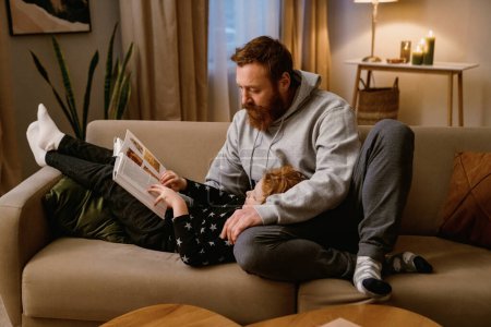 Foto de Padre e hijo pequeño leyendo libro mientras descansan en el sofá juntos en la acogedora sala de estar en casa - Imagen libre de derechos