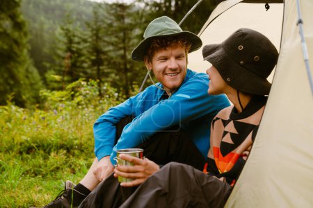 Photo for Young cheerful multiethnic tourist couple wearing panama hats drinking tea while sitting at camping tent in green forest - Royalty Free Image