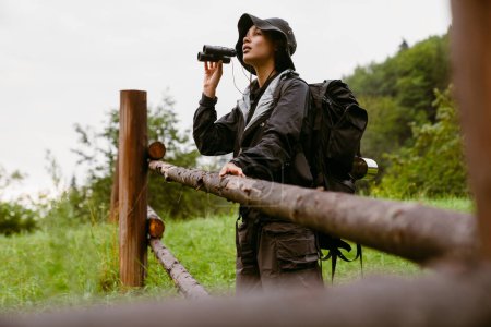 Photo for Young african woman tourist with backpack using binoculars standing near wooden fence during hiking in countryside - Royalty Free Image