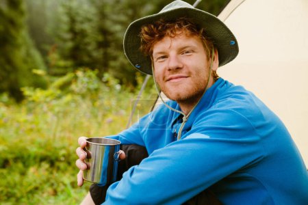 Photo for Young white tourist man drinking tea from metal cup while sitting at tent in forest - Royalty Free Image