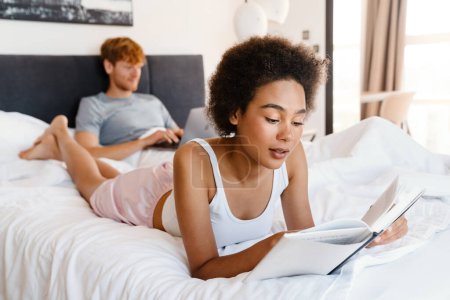 Téléchargez les photos : Jeune belle africaine bouclée femme lecture livre couché avec son petit ami qui travaille avec ordinateur portable sur le lit à la maison - en image libre de droit