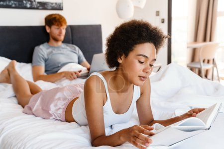Photo for Young beautiful african curly woman reading book lying with her boyfriend who working with laptop on bed at home - Royalty Free Image