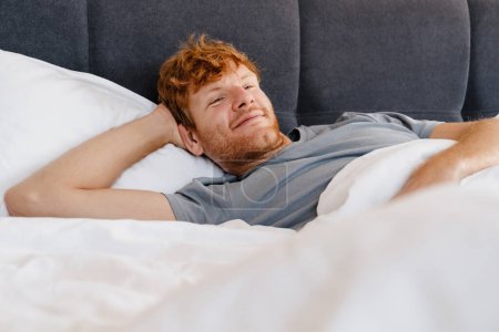Téléchargez les photos : Jeune homme rousse souriant beau en t-shirt gris avec la main derrière la tête regardant de côté allongé sur le lit à la maison - en image libre de droit