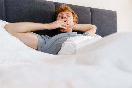 Foto de Joven hombre pelirrojo guapo en camiseta gris bostezando con los ojos cerrados acostado en la cama en casa - Imagen libre de derechos