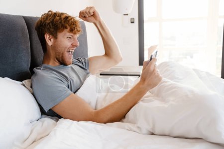 Téléchargez les photos : Jeune homme rousse beau et enthousiaste avec la bouche ouverte en t-shirt gris tenant le téléphone et faisant le geste du gagnant, tout en étant couché sur le lit à la maison - en image libre de droit