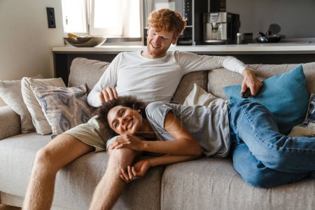 Photo for Young handsome smiling redhead guy caressing his beautiful african girlfriend liying on his knee, while they watching tv on couch at home together - Royalty Free Image