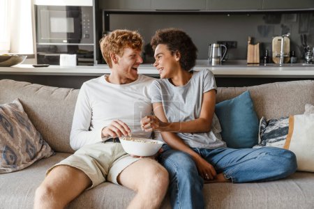 Photo for Young beautiful happy interracial couple eating popcorn and looking at each other, while sitting on couch in cozy sunny living room at home - Royalty Free Image