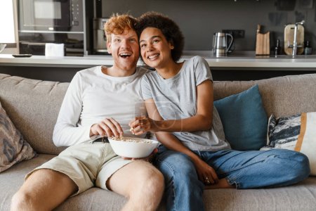 Téléchargez les photos : Jeune beau couple interracial heureux regarder le cinéma et manger du pop-corn, tout en étant assis sur le canapé dans le salon confortable et ensoleillé à la maison - en image libre de droit