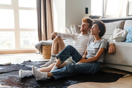 Photo for Young beautiful happy smiling interracial couple watching tv , while sitting on the floor leaning on couch at home together. Handsome redhead man and beautiful smiling african woman - Royalty Free Image