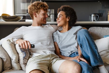 Photo for Young beautiful happy interracial couple hugging and looking at each other , while sitting on couch at home together. Handsome redhead man and beautiful smiling african woman - Royalty Free Image