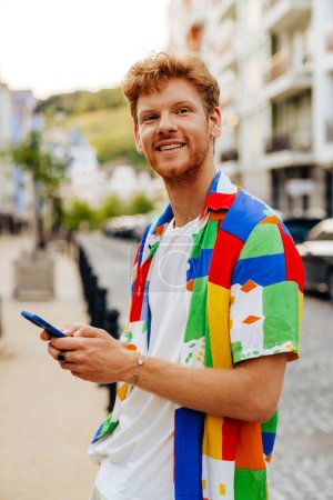 Téléchargez les photos : Jeune homme rousse souriant beau avec du chaume dans une chemise colorée tenant son téléphone et regardant de côté, tout en se tenant dans la rue - en image libre de droit