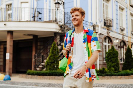 Foto de Joven pelirroja guapo sonriente hombre con rastrojo en colorida camisa caminando calle, cerca de edificio moderno - Imagen libre de derechos
