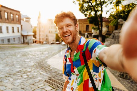 Téléchargez les photos : Selfie de jeune beau rousse souriant homme en chemise colorée debout dans la rue - en image libre de droit